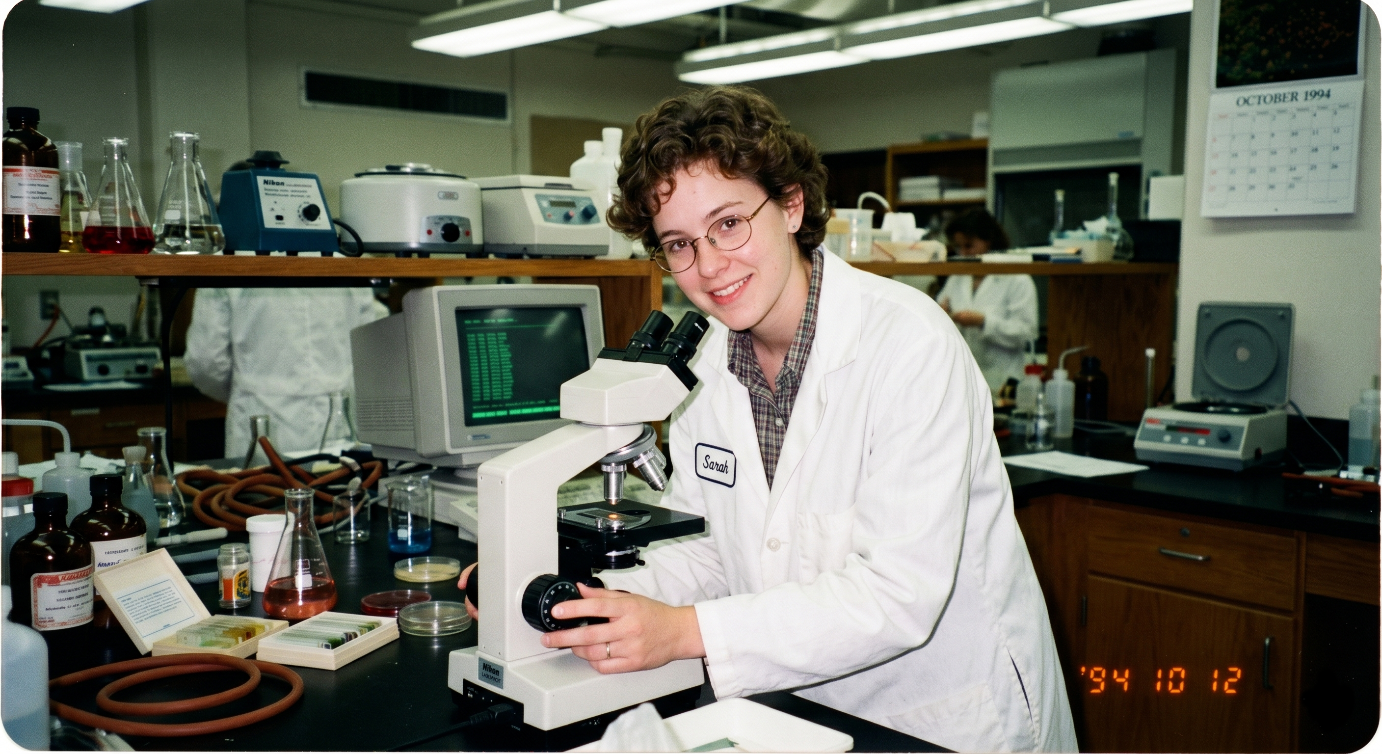 A young female scientist in a white coat smiling at a microscope in a laboratory, October 1994